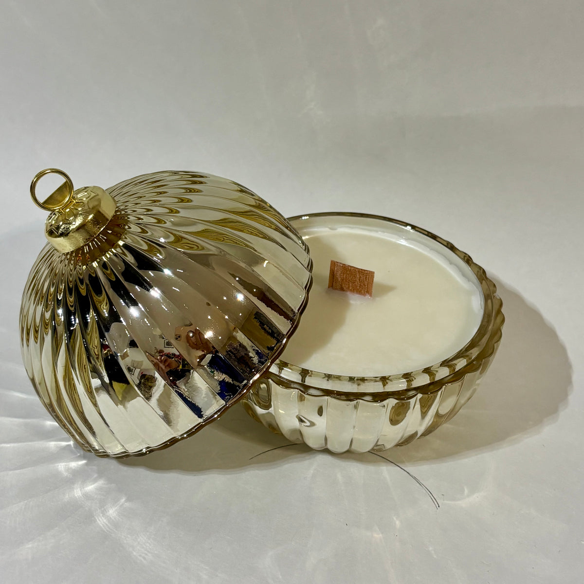 A glass jar candle with a ribbed, fluted design and a matching domed lid, shown open on a white background. The candle wax is white, and the lid and rim have a metallic gold finish.