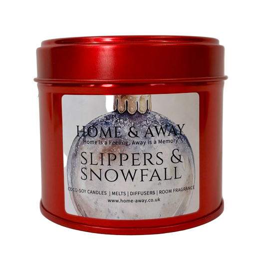 A red metal tin candle with a white label reading 'HOME & AWAY' and 'SLIPPERS & SNOWFALL' on a light neutral background.