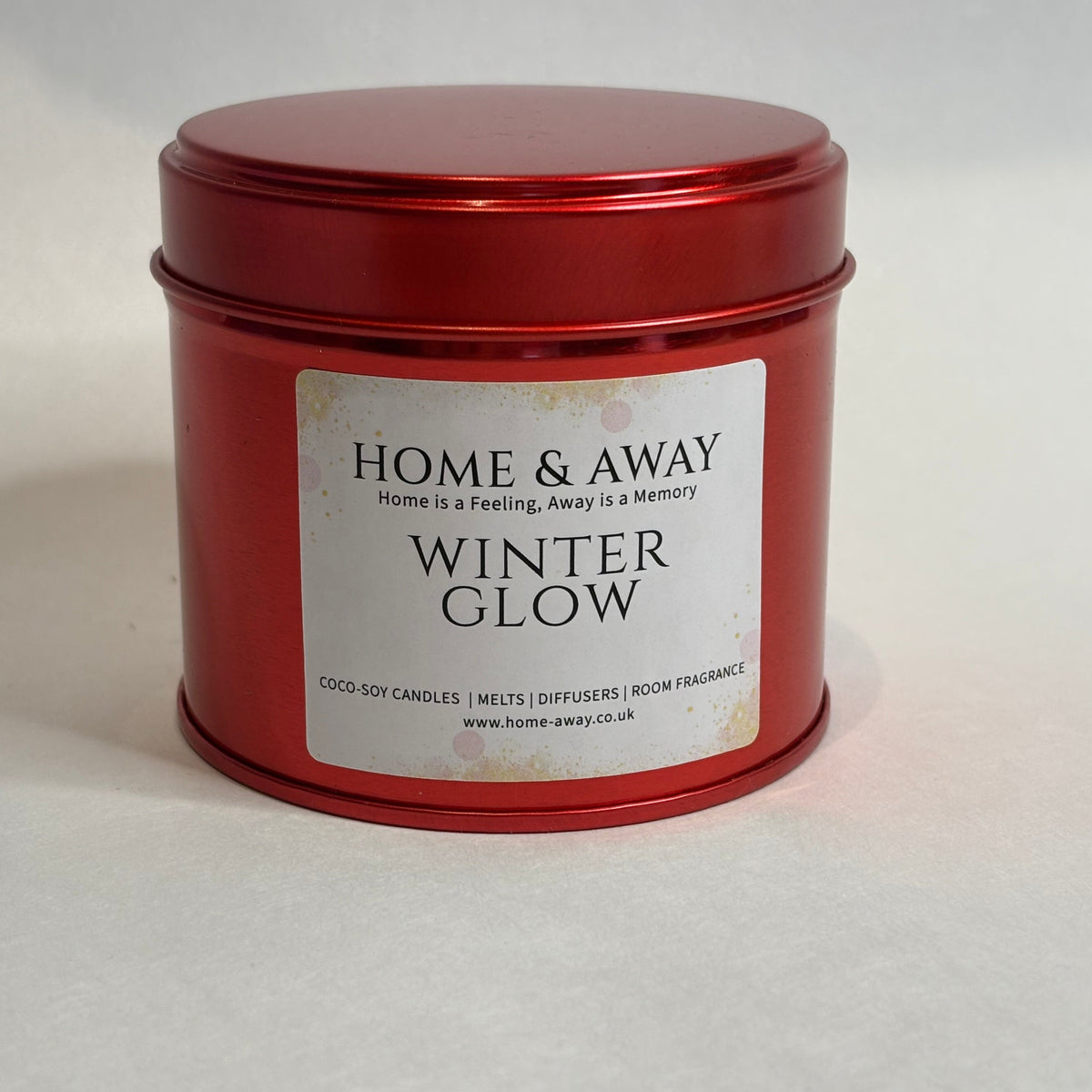 A red metal tin candle with a white label reading 'HOME & AWAY WINTER GLOW' on a light gray background.