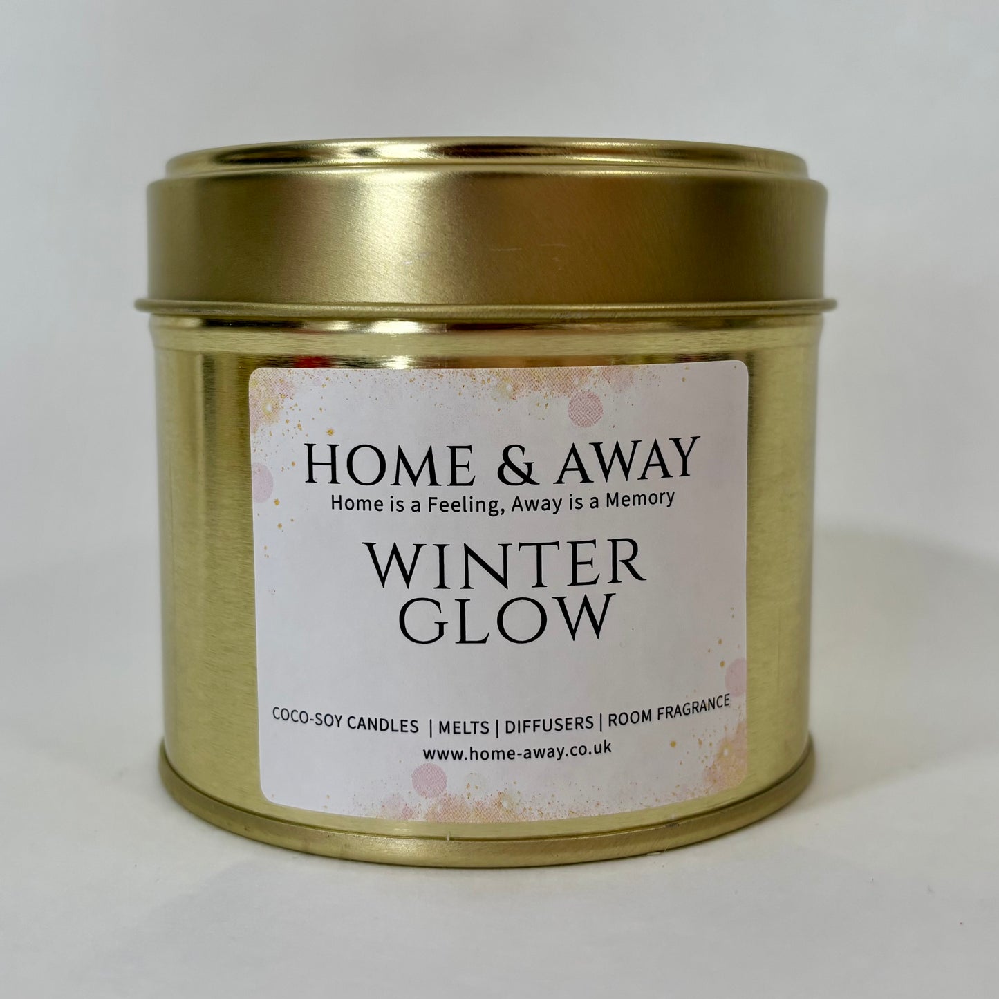 A gold metal tin candle with a white label reading 'HOME & AWAY WINTER GLOW' on a plain light background.