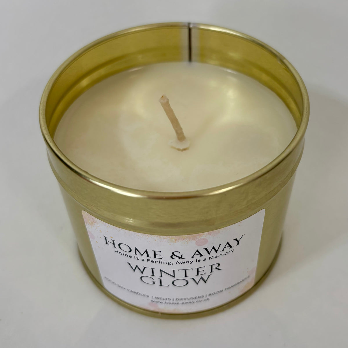 A single-wick soy candle in a round metallic gold tin with a white label reading 'HOME & AWAY WINTER GLOW' on a light neutral background.