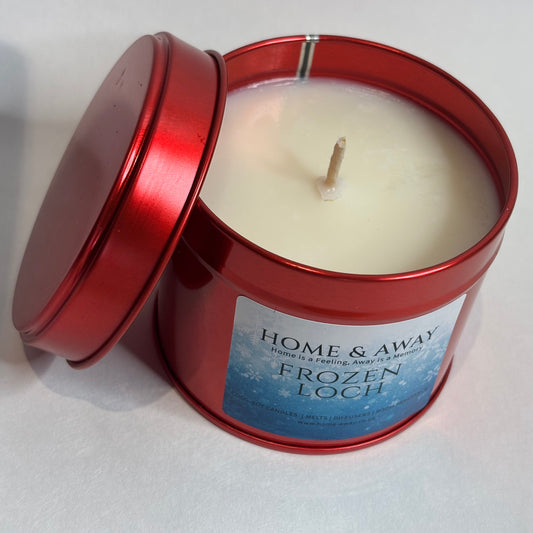 A red metal tin candle with its lid propped open, showing off-white wax and a single wick. The front label reads 'HOME & AWAY' and 'FROZEN LOCH'.