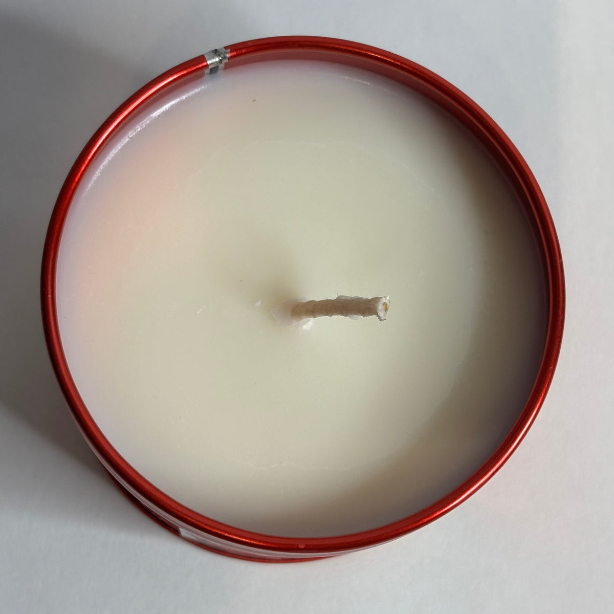 Top-down view of a single-wick candle in a round red metal tin, filled with creamy off-white wax, on a plain light background.