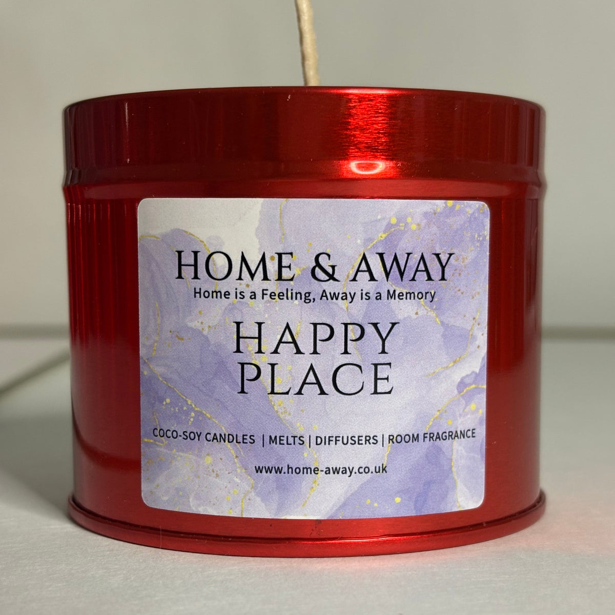 A red metal candle tin with a white wick visible at the top, featuring a front label that reads 'HOME & AWAY' and 'HAPPY PLACE' on a light purple marbled background.