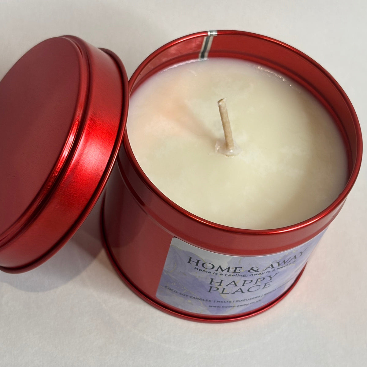 An open red metal tin candle with cream-colored wax and a single wick, shown on a light neutral background. The label reads 'HOME & AWAY' and 'HAPPY PLACE'.