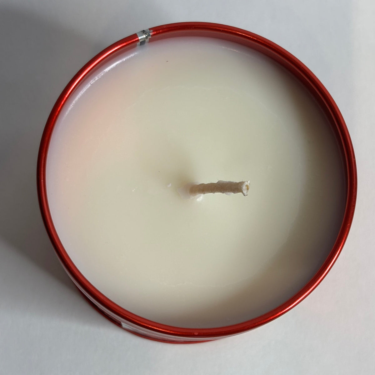 A top-down view of a single-wick candle in a round red metal tin, filled with off-white wax, set against a plain light gray background.