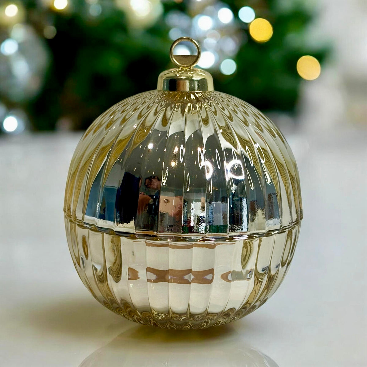 A round, ribbed glass Christmas ornament with a metallic gold finish, featuring a top hanging loop and a horizontal seam near the middle, set against a blurred festive background.