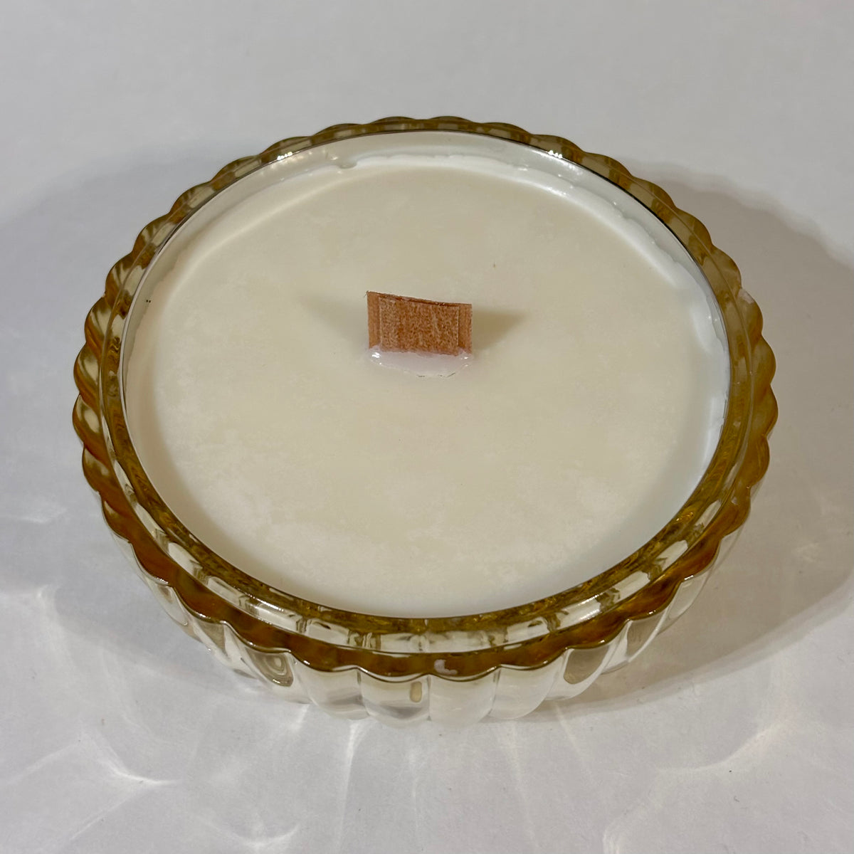 A round glass jar candle with a scalloped amber-brown rim and a single wick, filled with white wax, photographed on a light neutral background.