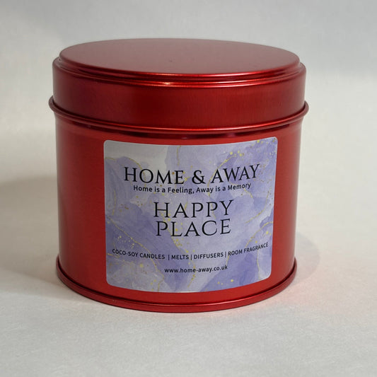A red metal tin candle with a matching lid, featuring a lavender and white label that reads 'HOME & AWAY HAPPY PLACE'.