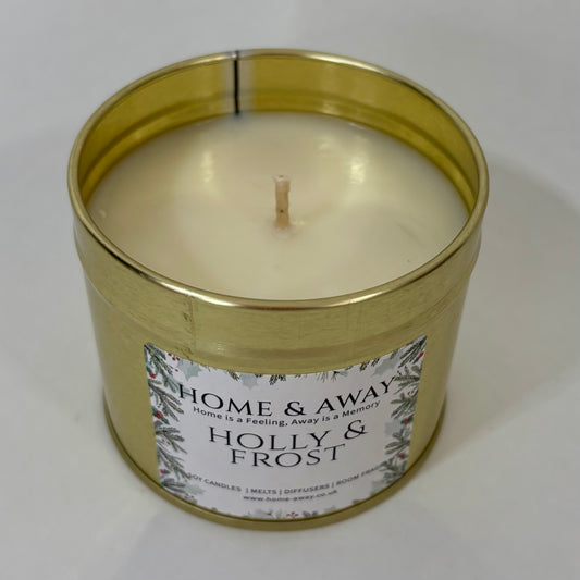 A gold metal tin candle with cream-colored wax and a white label reading 'HOME & AWAY' and 'HOLLY & FROST', photographed on a plain light background.